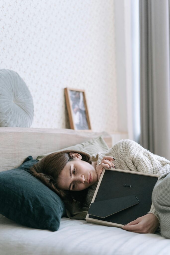 A woman reclines on a bed, reflecting with a picture frame in hand.